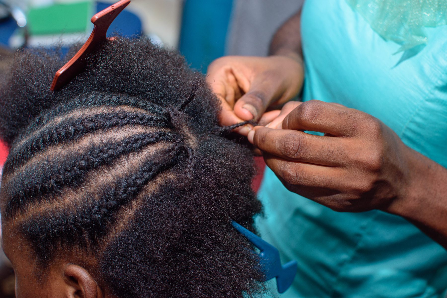Barber braiding client's hair with red comb, wearing turquoise shirt